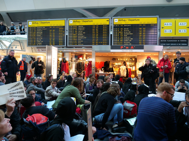 Eine große Gruppe von Menschen sitzt und steht in einem Flughafen während einer Protestaktion, mit Schildern, Puppen in Kleidern und Deckenleuchten im Hintergrund.