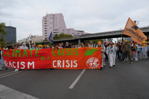 Eine Gruppe von Menschen marschiert eine baumbestandene Straße entlang und hält ein Banner mit der Aufschrift "Klimakrise ist eine Krise". Im Hintergrund sind Gebäude und eine Brücke zu sehen.