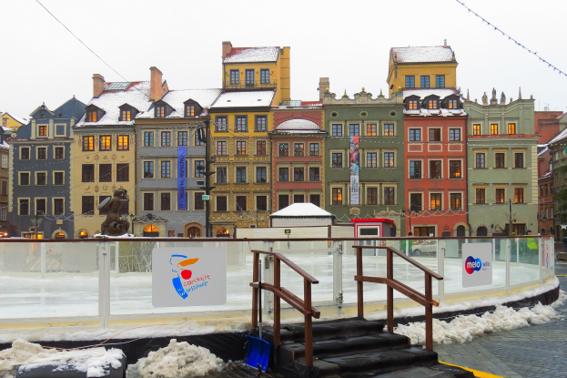 Eislaufbahn in einem Stadtplatz umgeben von Geb├Ąuden, Treppen, Gel├Ąndern, Informationsschildern, schneebedecktem Boden, Laternenmasten, B├Ąumen und Himmel.
