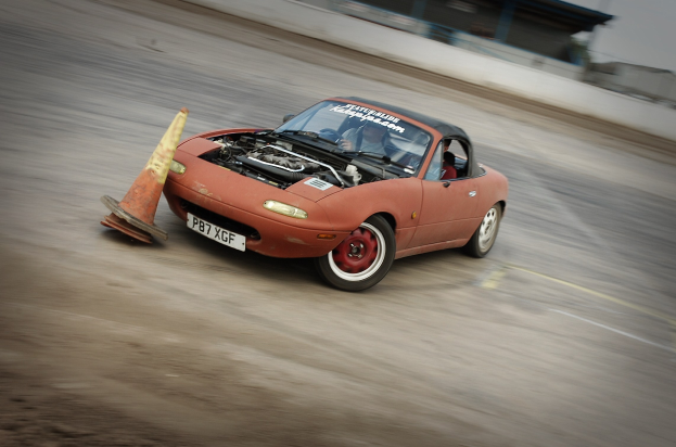 Eine Person, die ein rotes Auto auf einer Rennstrecke fährt, mit einem Verkehrskegel vorneweg, Hintergrund unscharf, was auf hohe Geschwindigkeit hindeutet.