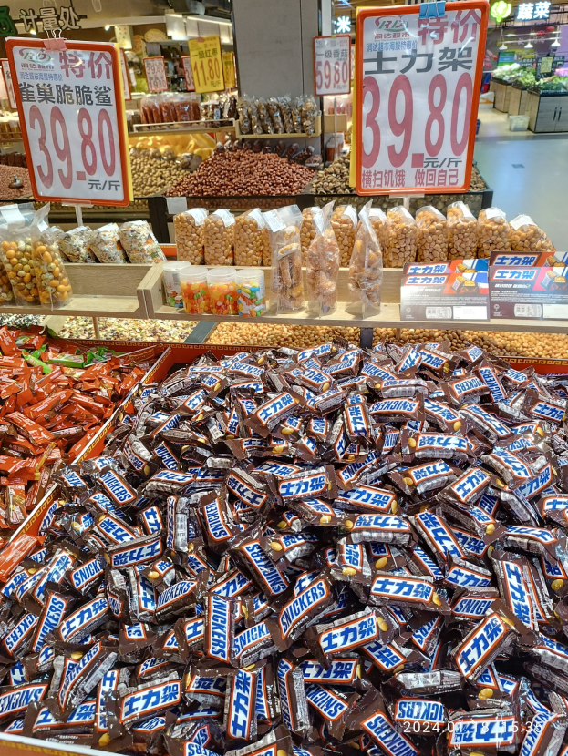 Eine Vitrine voller verschiedener Süßigkeiten, darunter Snickers-Riegel, in einem Geschäft mit Lebensmitteln auf Regalen und Deckenlampen im Hintergrund.