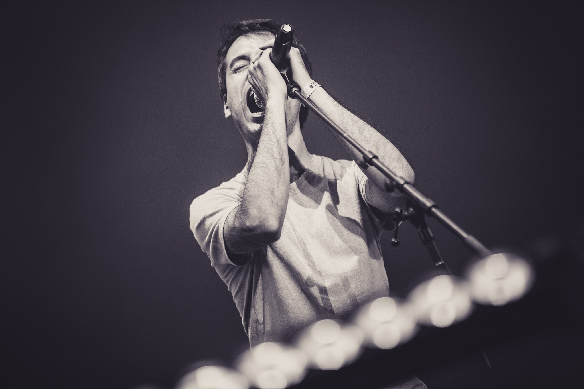 Ein Mann singt in ein Mikrofon auf einer hell erleuchteten Bühne, trägt ein schwarzes T-Shirt und Jeans, hat unordentliches Haar und schließt die Augen.
