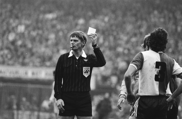 Schwarzes und weißes Bild eines Schiedsrichters mit roter Karte vor einer Menge in Fußballtrikots, mit einem Zaun im Hintergrund.