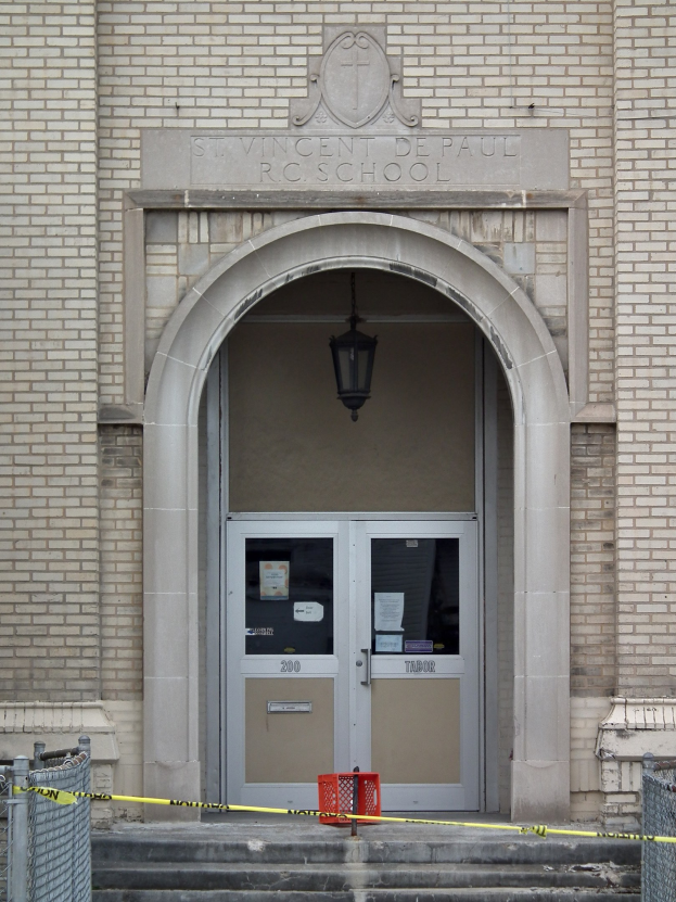 Der Eingang eines Backsteingebäudes mit gelbem Absperrband, Treppen mit Maschendrahtzaun, eine Tür mit Papieren, eine hängende Lampe und eine Wandinschrift, die wahrscheinlich den Namen der Schule angibt.