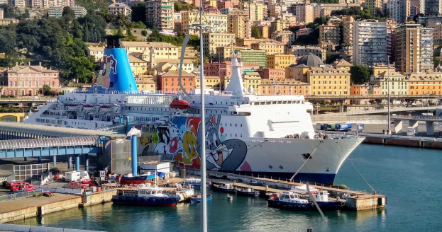 Großes Kreuzfahrtschiff an einem Stadtpier mit einer Brücke links, Fahrzeugen auf der Straße und Gebäuden und Bäumen im Hintergrund.