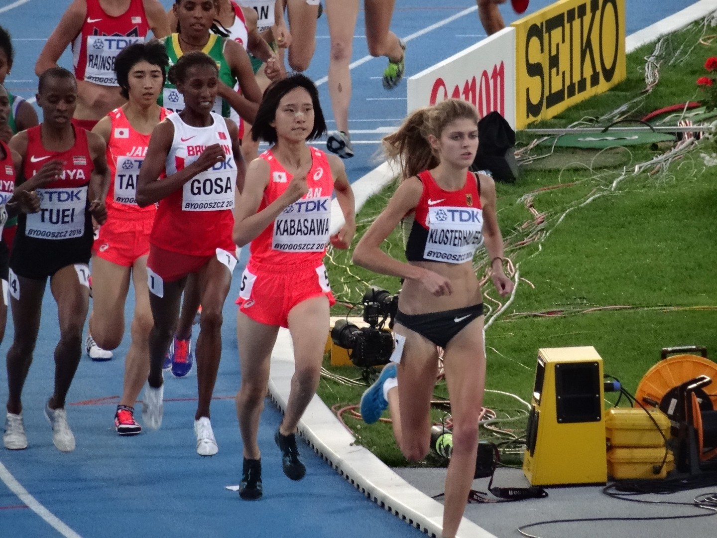 Gruppe von Frauen auf einer Laufbahn mit Kameramann im Vordergrund, Gras und Equipment rechts und farbenfrohe Blumen im Hintergrund.