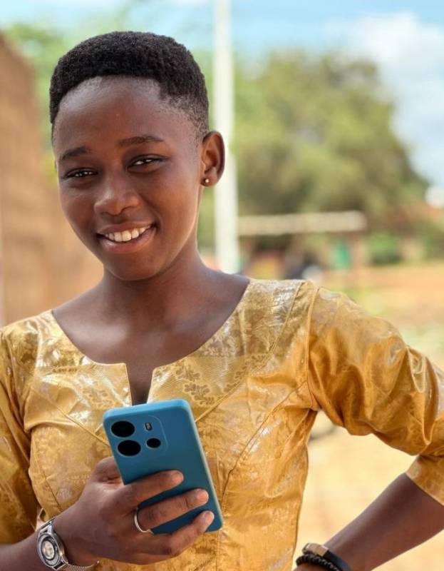 Ein junges Mädchen in einem gelben Kleid hält ein blaues Handy, lächelt vor einer Wand, einem Pfahl, Bäumen und Himmel.