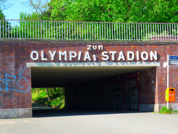 Der Eingang zum Olympiastadion in Berlin, Deutschland, mit einer Brücke mit Text, einem Metallzaun, einem Schild, einer Box, Pflanzen, Gras, einer Gruppe von Bäumen und einem bewölkten Himmel.