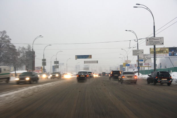 Eine städtische Straße mit starkem Verkehr an einem verschneiten Tag, mit Fahrzeugen, schneebedeckter Straße, Laternen, Texttafeln, Bäumen, Gebäuden und einem Himmel im Hintergrund.