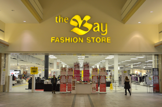 Außenansicht eines Bay Fashion Stores in einem Einkaufszentrum mit Menschen, Texttafeln an Wänden, Säulen, Deckenleuchten und Hintergrundobjekte.