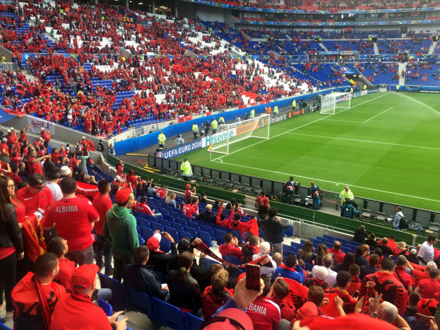 Eine große Menge Menschen in einem Stadion schaut ein Fußballspiel, mit Menschen auf Stühlen und Stehplätzen, Schildern, einem Tor und Deckenleuchten.