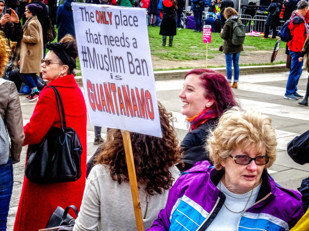 Eine Gruppe von Menschen steht im Freien, einige halten Taschen, eine Frau hält ein Schild mit der Aufschrift "Der einzige Ort, der ein Muslimverbot benötigt, ist Guantanamo", neben einem Metallzaun, einem Schild, einem Gebäude und Bäumen.