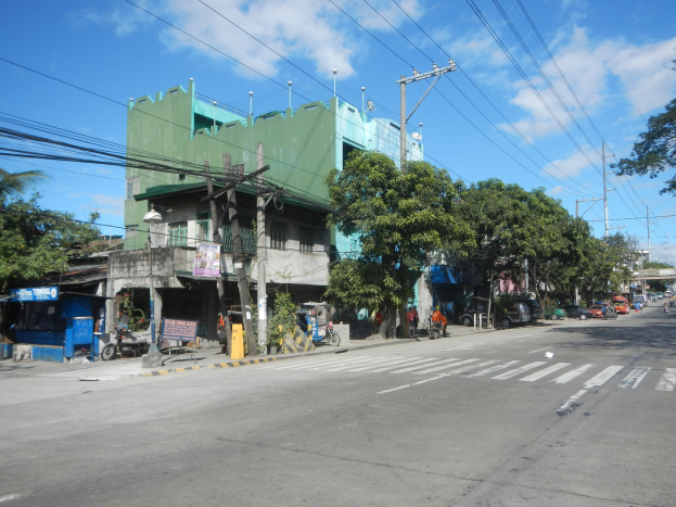 Eine Stadtstraße mit einem grünen Gebäude, fahrenden Fahrzeugen, Fußgängern auf dem Gehweg, Strommasten mit Drähten und einem bewölkten Himmel.