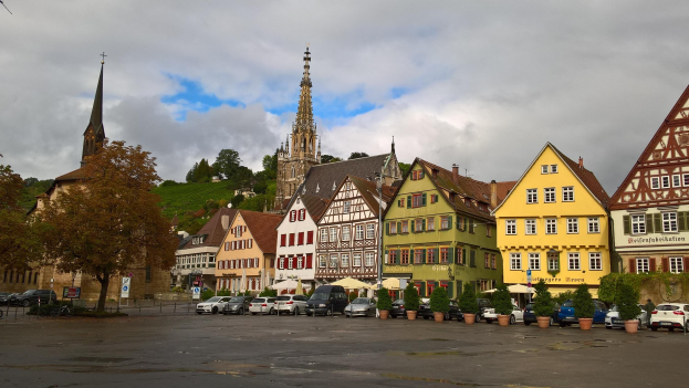 Ein malerischer Marktplatz in Rothenburg ob der Tauber, Deutschland, mit Gebäuden, Bäumen, Topfpflanzen, Fahrzeugen, Schildern und einer Kirche unter einem bewölkten Himmel.