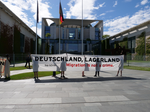 Eine Gruppe von Menschen hält eine Fahne mit der Aufschrift "Deutschland, Lagerland Migration ist kein Verbrechen" vor einem umzäunten Gebäude mit Bäumen und Flaggen.