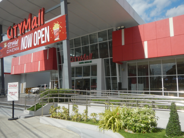 Stadtmalleingangsschild mit der Aufschrift "Stadt Mall Now Open" mit umgebender Vegetation, Menschen, einem Auto, Pfählen, einer Fahne, Lichtern und einem bewölkten Himmel.