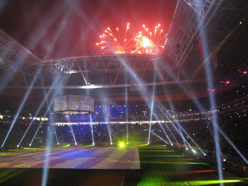 Olympiastadion in London bei Nacht, erleuchtet mit hellen Lichtern und Feuerwerk, umgeben von einer Menge und einem großen Bildschirm im Vordergrund.