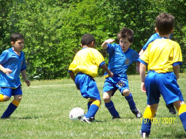 Kinder spielen Fußball auf einem grünen Rasen mit Bäumen im Hintergrund und einem Datumsstempel in der rechten unteren Ecke.