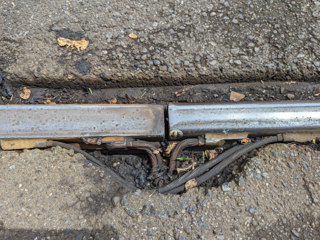 Nahaufnahme eines metallenen Kanals auf der Straße mit daran befestigten Drähten und verstreuten trockenen Blättern.