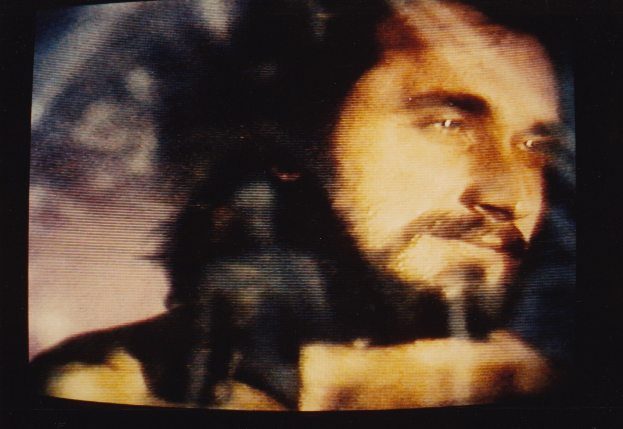 Ein Mann mit langem dunklem Bart und Haar, der ein weißes Hemd trägt, erscheint auf einem Fernsehbildschirm mit einem ernsten Gesichtsausdruck, seine Augen und leicht geöffneten Lippen sind auf den Betrachter gerichtet, vor einem dunklen Hintergrund.