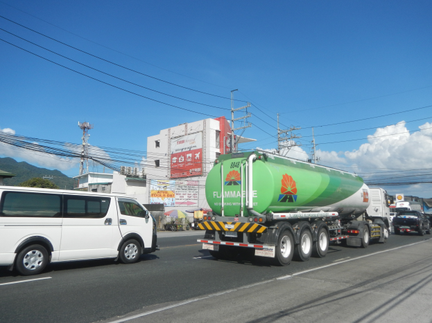 Ein gro├če Tanklastwagen fahrt auf einer Stra├če neben einem wei├če Van, mit Strommasten, Geb├Ąuden, Plakaten, B├Ąumen, H├Ąngen und einem bew├Âlktem Himmel im Hintergrund.