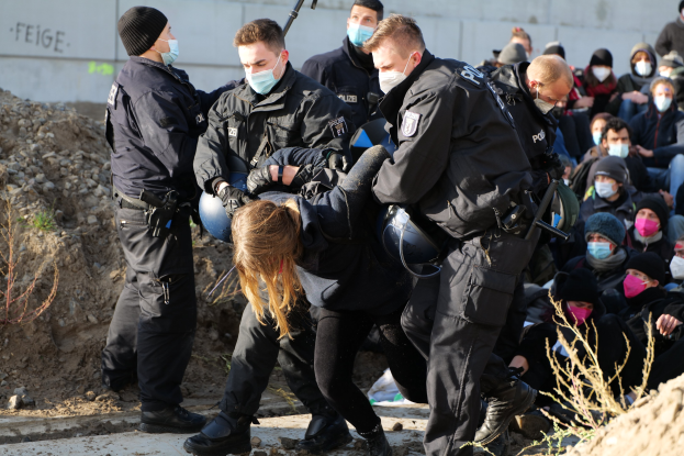Gruppe maskierter Personen in Mützen, einige bewaffnet, umringt eine Frau, die von der Polizei eskortiert wird, vor einer Wand mit Text und städtischer Vegetation, was auf eine Protestaktion gegen Regierungssparmaßnahmen hinweist.