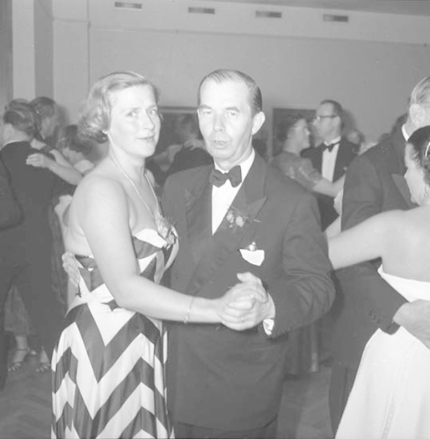 Ein Mann in einem schwarzen Blazer, weißem Hemd und schwarzen Hosen tanzt mit einer Frau in einem unbekannten Outfit auf einer formellen Veranstaltung, indem sie sich an den Händen halten und zur Musik schwenken, vor einem Wandhintergrund.