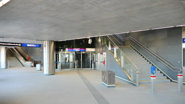 Leerer Bahnhof mit Treppe, Geländern, Mülltonnen, Texttafeln, Deckenleuchten und Glastüren im Hintergrund.