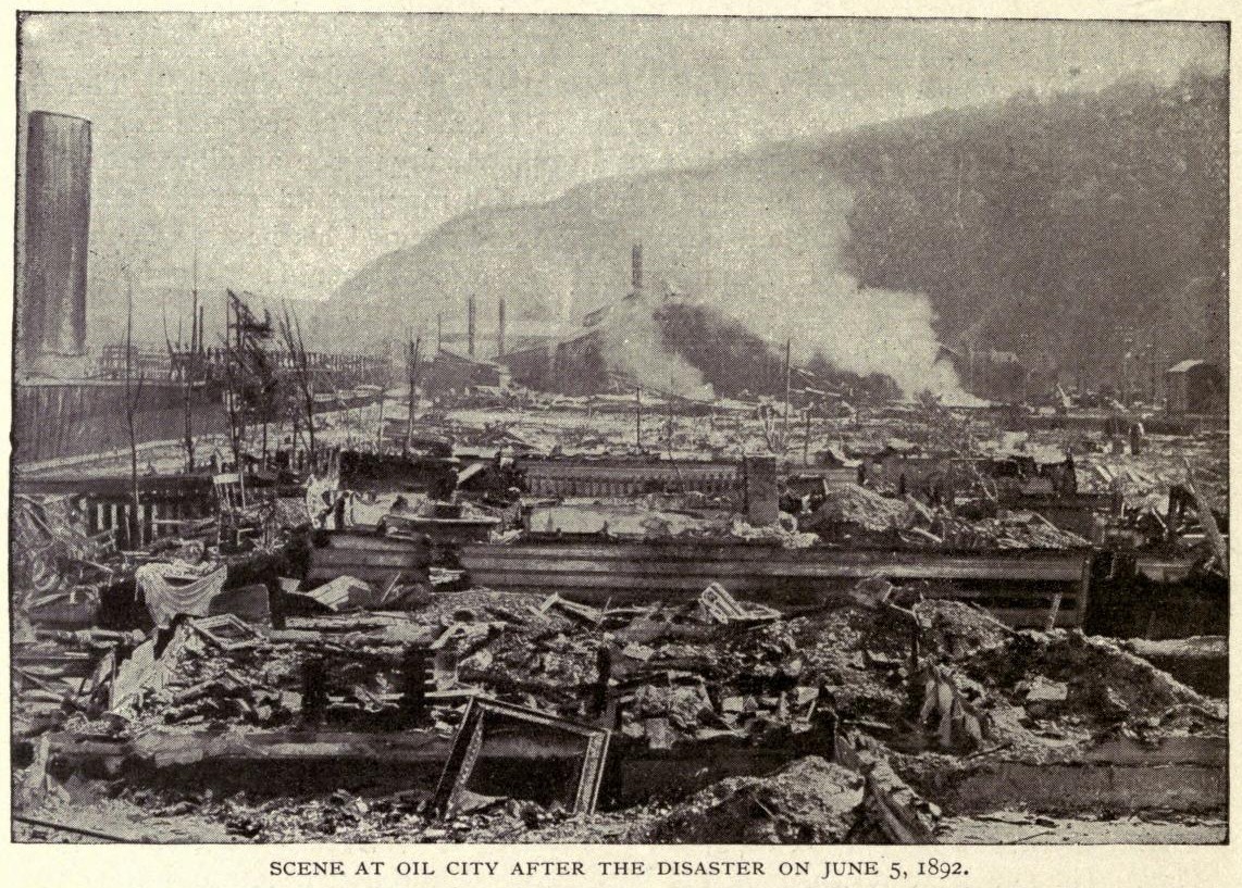 Ein Schwarz-Weiß-Foto einer Stadt nach einer Katastrophe am 5. Juni 1892, das beschädigte Gebäude, Pfähle, Rauch und einen Hügel im Hintergrund zeigt, mit Text am unteren Rand.