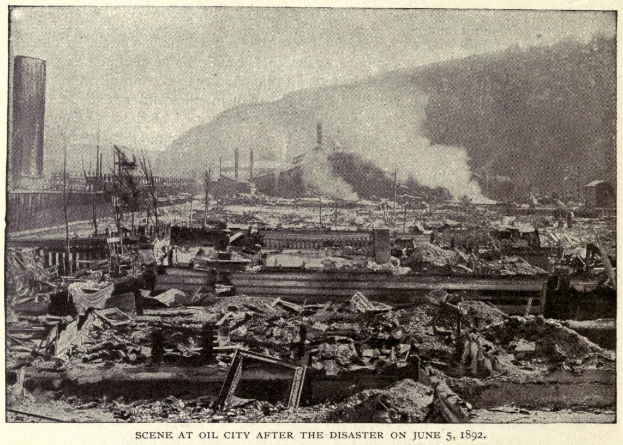 Ein Schwarz-Weiß-Foto einer Stadt nach einer Katastrophe am 5. Juni 1892, das beschädigte Gebäude, Pfähle, Rauch und einen Hügel im Hintergrund zeigt, mit Text am unteren Rand.