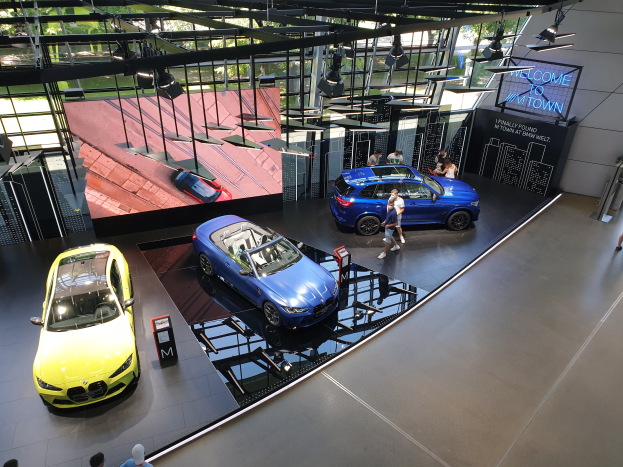 Eine Gruppe von Menschen im BMW Museum in München, mit Ausstellungsautos links und einem Schild mit der Aufschrift "BMW Museum München" rechts, mit Blick auf eine städtische Außenansicht durch Glasfenster.