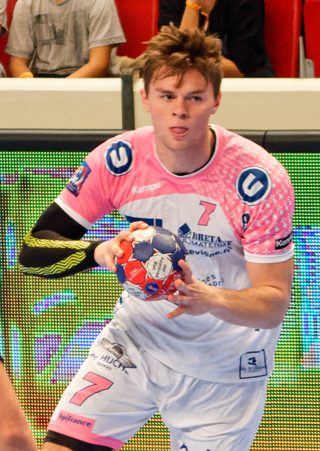 Ein Mann in einem pinken Hemd hält einen Fußball, wobei die Hand einer anderen Person auf der linken Seite zu sehen ist und im Hintergrund einige Menschen auf Stühlen sitzen.