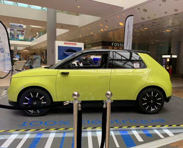 Gelbes wasserstoffbetriebenes Elektroauto vor einem Gebäude mit Absperrpollern, Seilen, Banner, Personen, Schildern, einer Topfpflanze, Deckenleuchten und Ventilatoren.