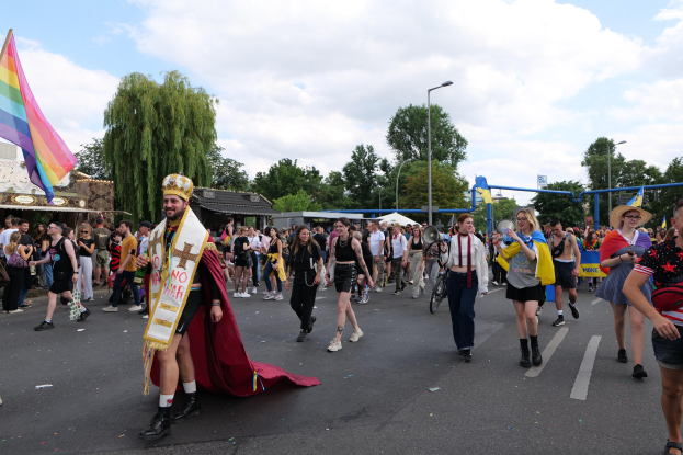 Eine Gruppe von Menschen bei der Gay Pride Parade 2018 mit einer Regenbogenfahne und Musikinstrumenten, umgeben von Laternenmasten, Bäumen und Hütten.
