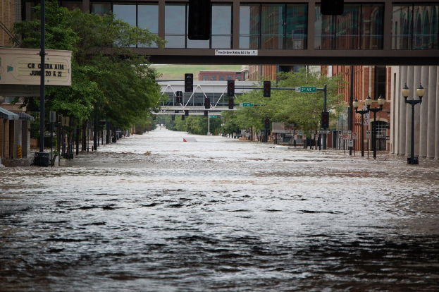 Vollständig überflutete Straße mit Wasser auf der Straße, an Masten, Laternen, Verkehrsampeln, Bäumen, Gebäuden und einer Brücke im Hintergrund.