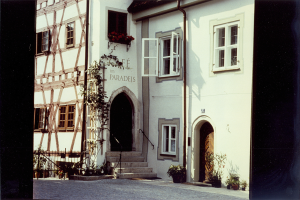 Ein altes Gebäude mit weißer Fassade und schwarzer Tür, umgeben von Zimmerpflanzen, Treppen, einem Geländer und einem Weg, mit mehreren Fenstern.