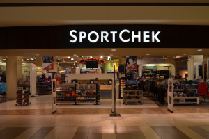 Außen- und Innenansicht eines Sportchek-Ladens in einem Einkaufszentrum mit Kleiderstangen, Deckenleuchten, Säulen und einem Fernseher.