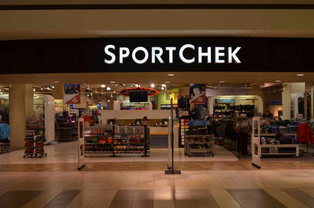 Außen- und Innenansicht eines Sportchek-Ladens in einem Einkaufszentrum mit Kleiderstangen, Deckenleuchten, Säulen und einem Fernseher.