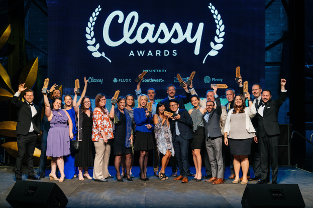 Gruppe von Menschen auf einer Bühne, die Auszeichnungen halten, mit Lautsprechern unten und einem Bildschirm im Hintergrund mit 'Classy Awards 2019'
