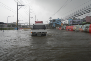 Ein Van fährt durch eine überflutete Stadtstraße mit Strommasten, Laternenmasten und Gebäuden auf beiden Seiten, eine Brücke im Hintergrund und der Himmel darüber sichtbar.