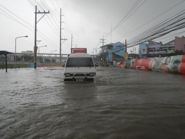 Ein Van fährt durch eine überflutete Stadtstraße mit Strommasten, Laternenmasten und Gebäuden auf beiden Seiten, eine Brücke im Hintergrund und der Himmel darüber sichtbar.