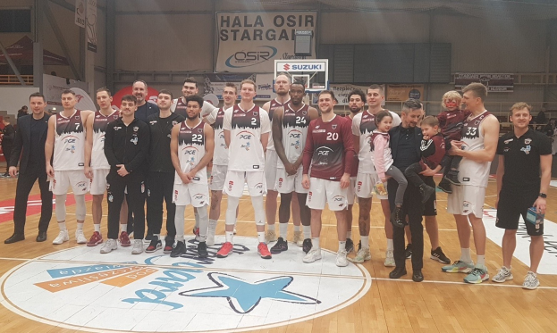 Männer- und Kinder-Gruppe auf einem Basketballfeld mit Schildern "Hala Osir Starga", einem Basketballkorb, sitzenden Zuschauern und Innenbeleuchtung.