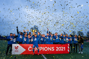 Eine Gruppe von Männern in blauen T-Shirts steht auf einem Fußballfeld und hält einen Pokal hoch, mit einem roten "Champions 2021"-Schild und fliegenden gelben Papieren im Hintergrund.