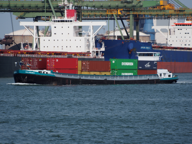 Ein großes Containerschiff voller Container liegt nahe einem Hafen mit einer Brücke im Hintergrund bei klarem Himmel, mit Text am unteren Bildrand.