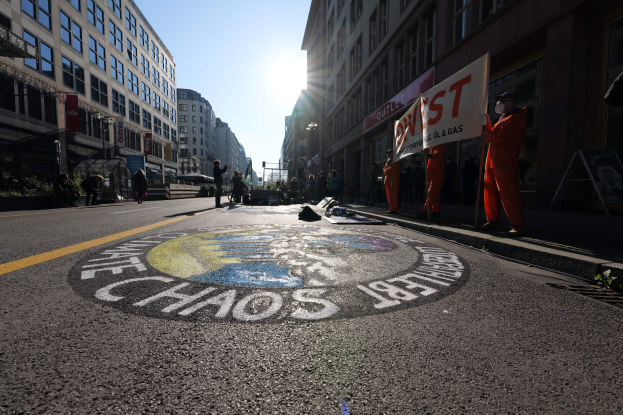 Menschen mit Schildern und Plakaten stehen auf einer Straße, die von Gebäuden und Pflanzen gesäumt ist, mit "Chaos" auf die Straße gemalt während einer Demonstration.