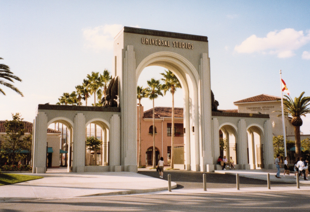 Eingang von Universal Studios Hollywood mit einem Bogen, einer Statue, Menschen, Bäumen, Gebäuden und einem Flaggenmast.