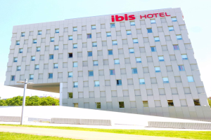 Das Ibis Hotel in Berlin, Deutschland, ein großes Gebäude mit Fenstern, steht unter einem bewölkten Himmel, mit einem Straßenschild, einer Gruppe von Bäumen, einem Zaun und einem Weg im Vordergrund.