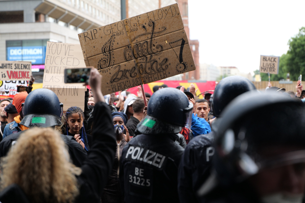 Gruppe von Menschen mit Helmen und Schildern vor einer Menge, mit Gebäuden, Bäumen und einem Pfahl im Hintergrund.