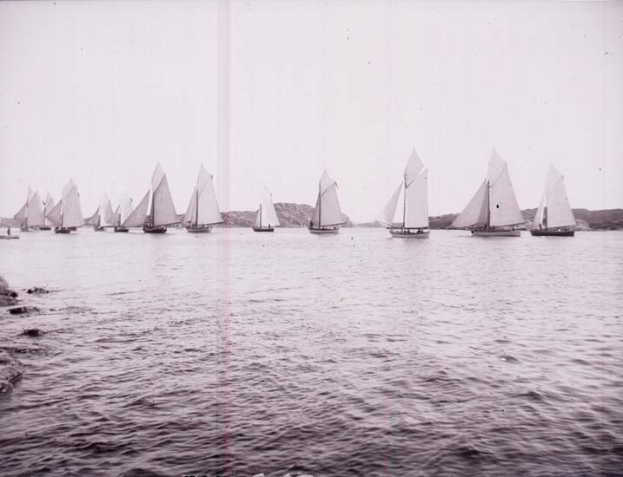 Schwarzes und weißes Foto von Segelbooten auf dem Wasser mit Hügeln im Hintergrund und einem klaren Himmel, als Foto des New York Yacht Club bezeichnet.
