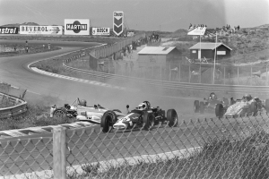Ein Schwarz-Weiß-Foto von Autos auf einer Rennstrecke, bei der die Fahrer Helme tragen, ein Maschendrahtzaun im Vordergrund und eine Hintergrundansicht mit Texttafeln, Schuppen, Pfosten, Menschen, Hügel und Himmel.